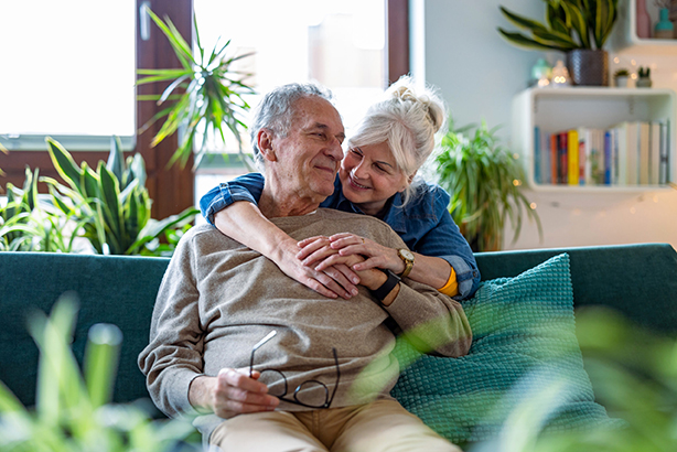 Elderly man on a sofa, wife behind with arms wrapped around him, looking at each other smiling.
