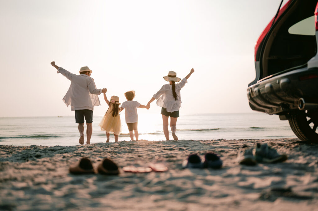 A family – mum, dad, girl and boy - running on the sand on a sunny day towards the sea, holding hands, arms in the air, having kicked off their shoes, with the car in the foreground